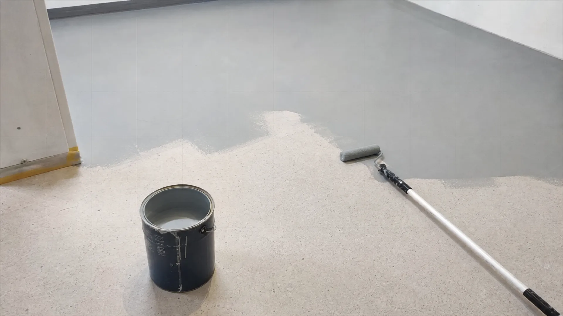 Ein Garagenboden wird mit grauer Farbe gestrichen, ein Farbeimer und eine Rolle liegen auf dem Boden.
